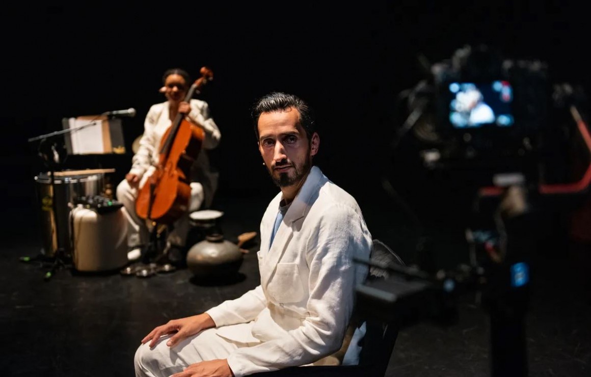 Rachif El Kaoui zit zijwaarts op stoel en kijkt recht in camera. Hij draagt een volledig wit pak. In de achtergrond zijn we een cellist zitten. 