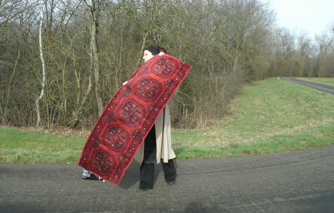 Emine Cerkinaj staat in een straatje, achter haar zien we een veld en bomen. Ze houdt een tapijt voor haar vast. 