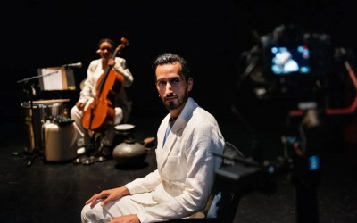 Rachif El Kaoui zit zijwaarts op stoel en kijkt recht in camera. Hij draagt een volledig wit pak. In de achtergrond zijn we een cellist zitten. 